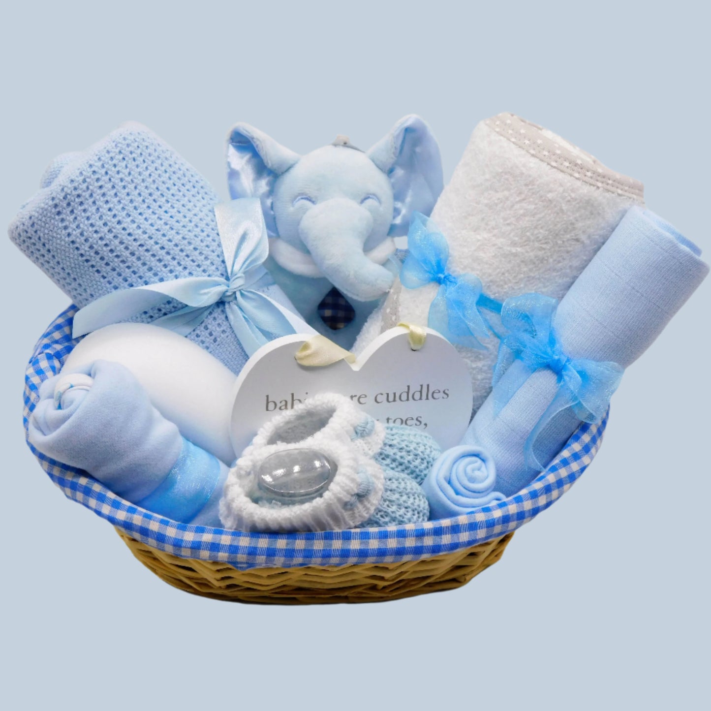 Baby Boy gift basket with blue and white items including a plush elephant rattle, blanket, towel, clothing and booties on a pale blue background.