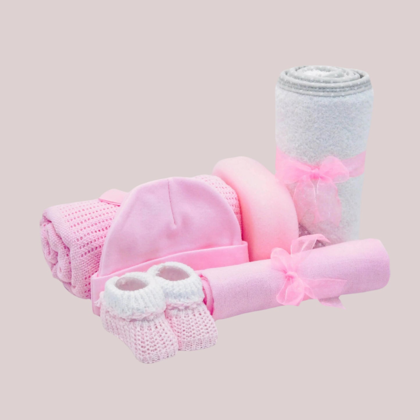Baby gift set with pink hat, booties, muslin square, sponge and blanket and white towel on a light pink background