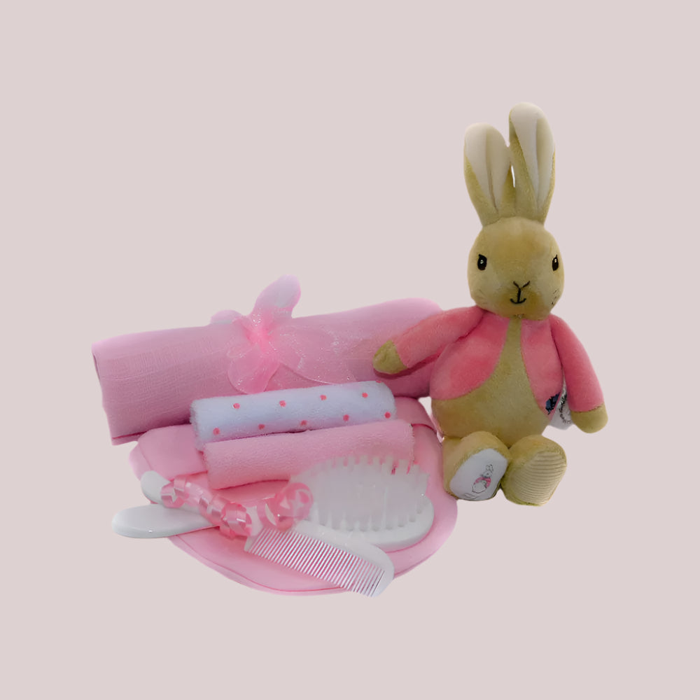 Set of baby items including a plush Flopsy Bunny bean rattle, muslin square, brush and comb and washcloths on a pale pink background