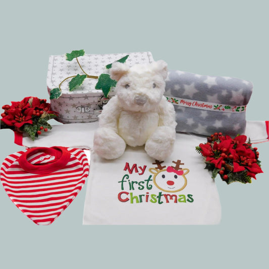 Christmas Baby gift set with white teddy bear, bib, blanket and sleepsuit with 'My first Christmas' text, surrounded by festive decorations.