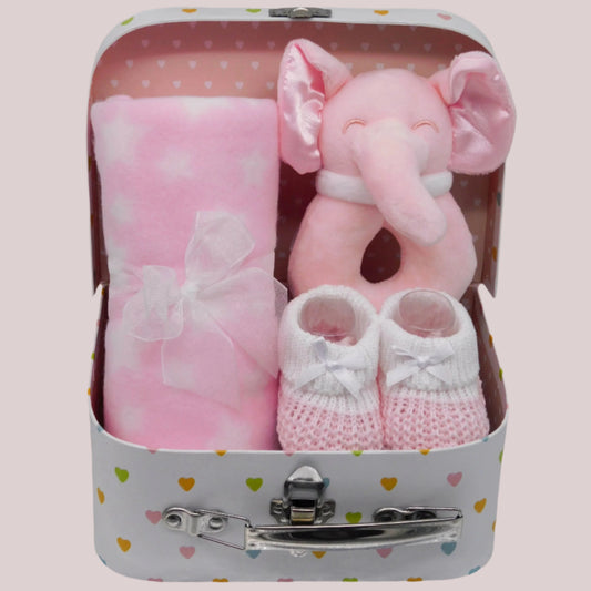 Baby gift set with plush pink elephant rattle, pink and white blanket, and knitted booties in a keepsake suitcase on a pale pink background