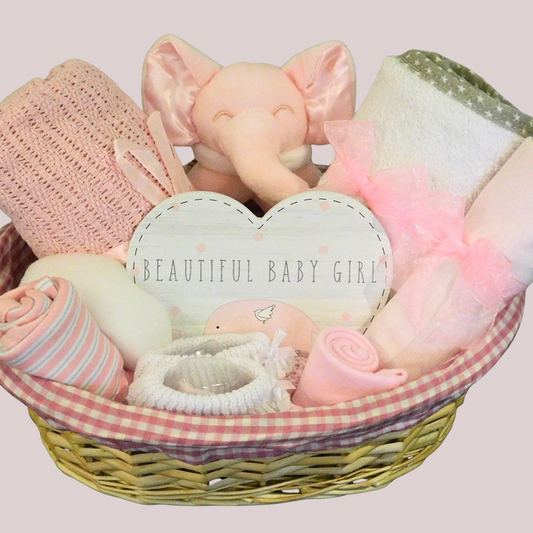Oval wicker baby gift basket with pink & white gingham lining, displaying towel, blanket, booties, hat, muslin, sponge. bodysuit, heart-shaped plaque & elephant rattle