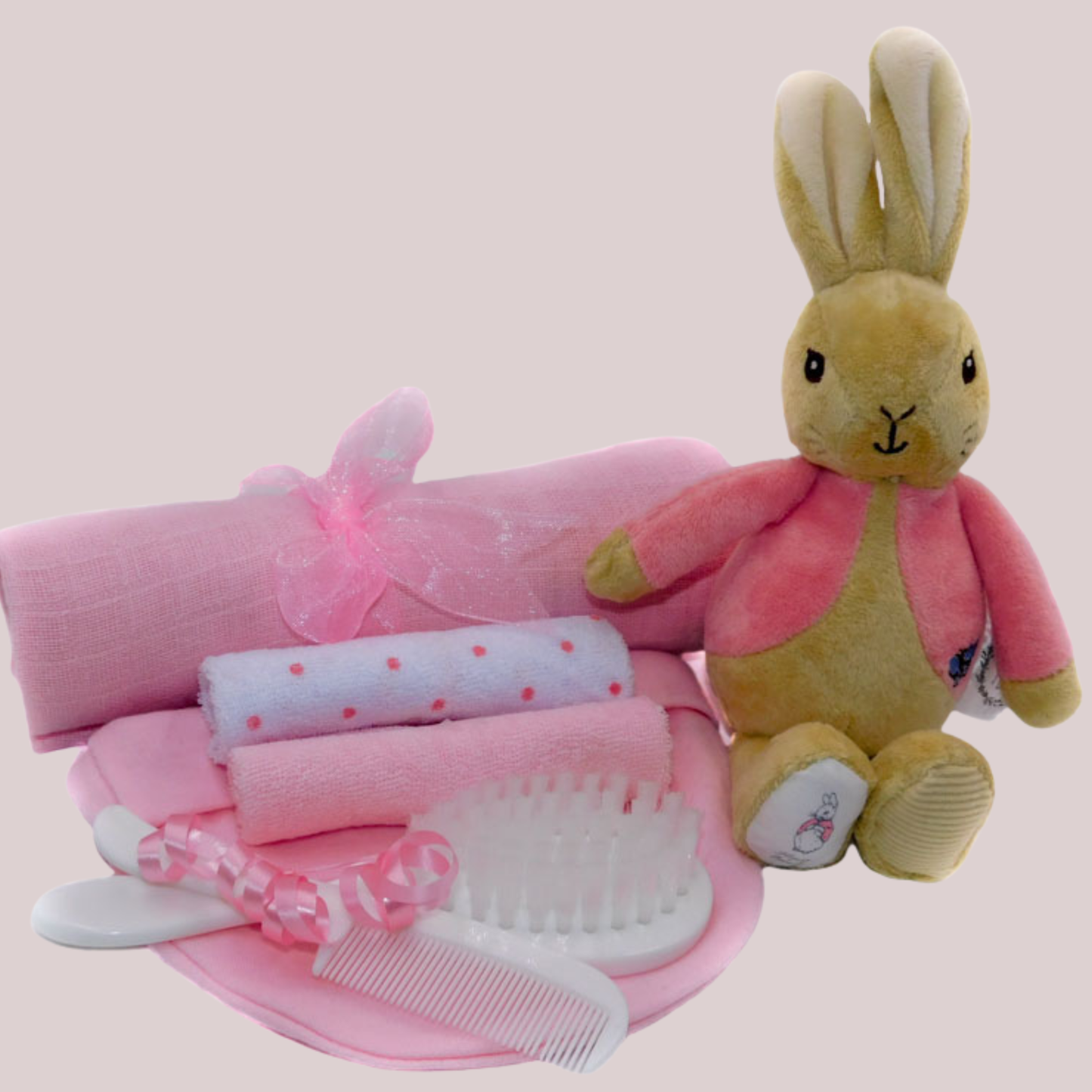 Set of baby items including a plush Flopsy Bunny bean rattle, muslin square, brush and comb and washcloths on a pale pink background