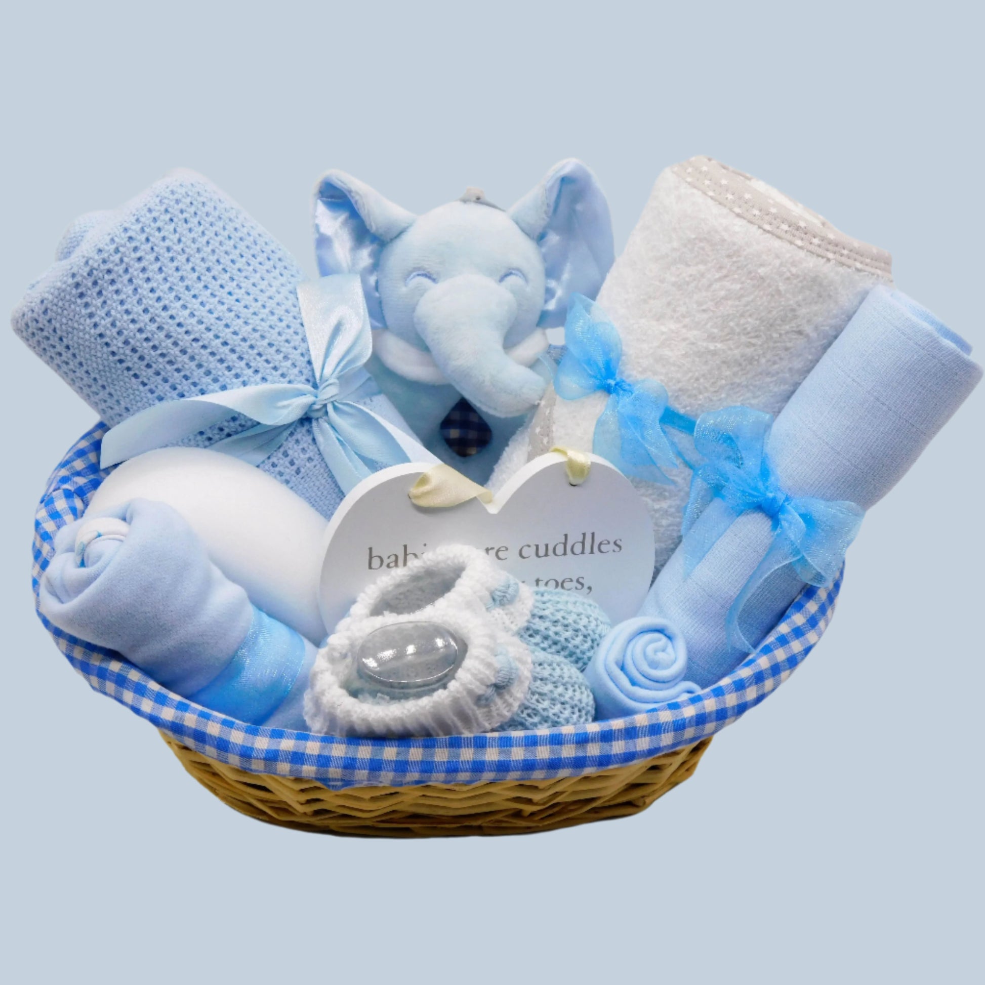 Baby Boy gift basket with blue and white items including a plush elephant rattle, blanket, towel, clothing and booties on a pale blue background.