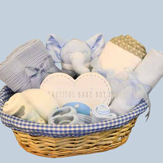 Wicker Baby gift basket with blue & white gingham lining, displaying baby towel, blanket, hat, booties, bodysuit, muslin, sponge, elephant rattle & heart-shaped plaque
