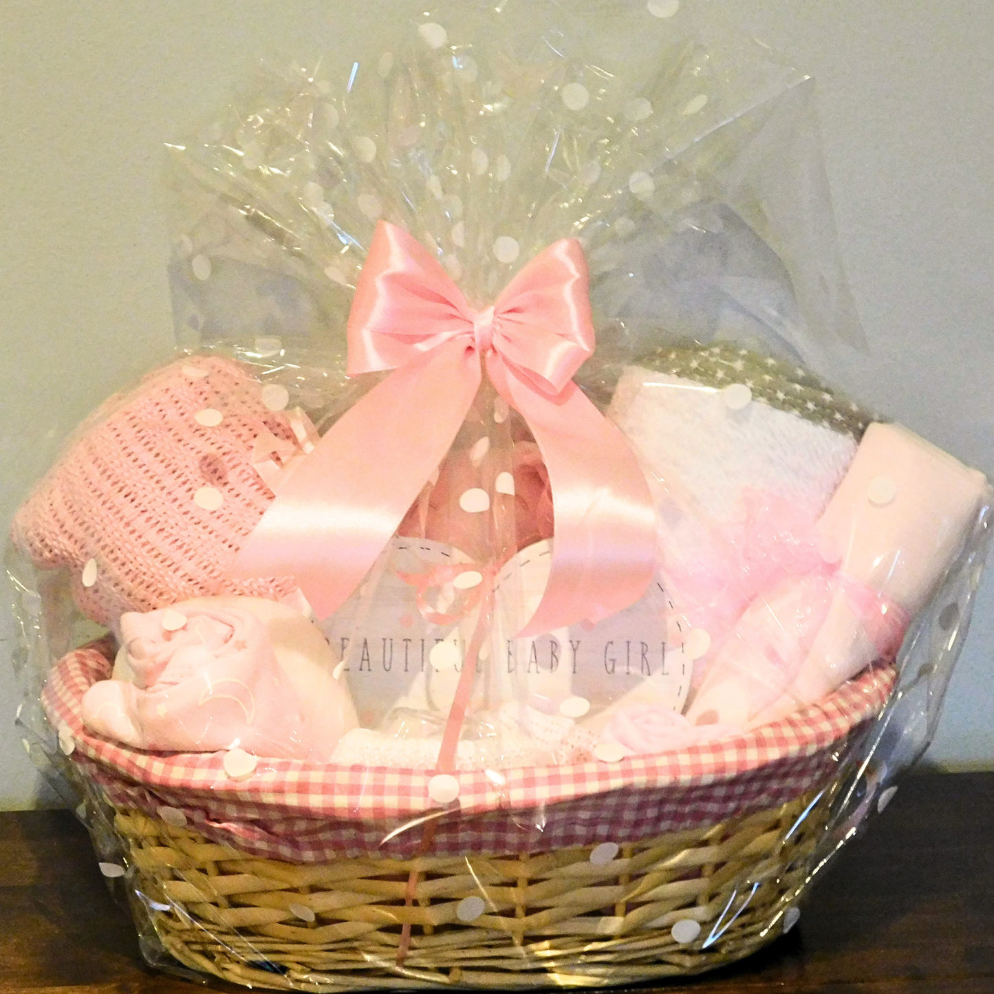 Baby gift basket, wicker with pink & white gingham lining, filled with baby essentials & keepsakes & wrapped in cellophane with a pink bow